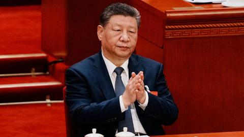 Chinese President Xi Jinping adjusts his navy blue jacket as he sits at the closing session of the Chinese People's Political Consultative Conference (CPPCC) at the Great Hall of the People on March 11, 2026 in Beijing, China. He is sitting in front of a long wooded table with two white tea cups in front of him.