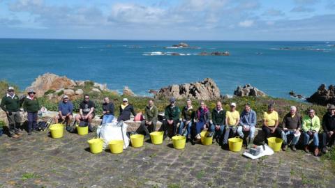 Image shows a large group of volunteers sitting on rocks, against a backdrop of the blue sea and coastline, with large yellow collection buckets in the foreground.