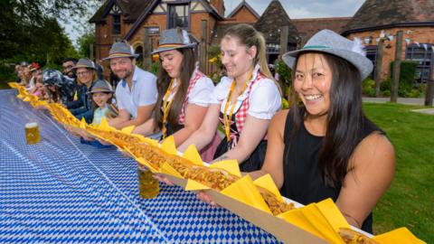 A line of people, some wearing grey hats, hold a very long sausage over an equally long table which has a blue and white table cloth over it.