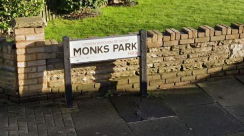 A Google street view of a road sign reading: Monks Park. It is on a pavement, in front of a grassy front lawn. 