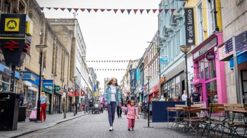 A woman dressed in purple leggings, a grey top and blue jacket is walking down a pedestrianised road holding hands with a young girl who is wearing pink trousers and a pink coat. Café tables and chairs are on the road and bunting crosses between the buildings. The pair are walking past a Liverpool Central Merseyrail station logo.