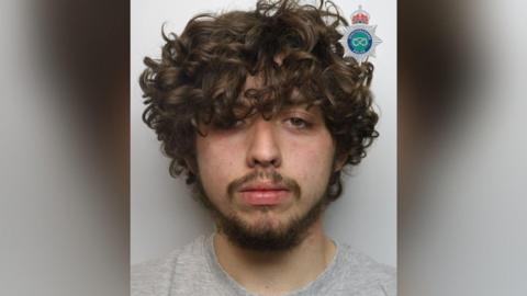 A mugshot of a man with brown curly hair, which is partially covering his eyes, with a black beard and a grey shirt.