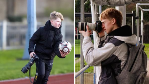 A boy (left) with blond hair is wearing a black coat and tracksuit bottoms. He is holding a camera with a long lens in one hand, and a football in the other. The second boy (right) is holding a camera with a long lens to his eye, while leaning his elbow on a pole. He is wearing a light grey and black jacket and grey rucksack.
