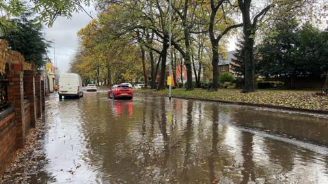 A flooded road which is lined with trees on one side and there is a brick wall on the other. There are vehicles driving through the water, with a red car in the middle of the image and a white van to the left hand side of the road along with other vehicles in the distance.