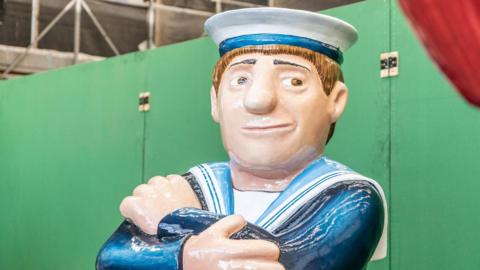 A sailor sculpture in the style of Beryl Cook's paintings sits with his arms crossed. He's wearing a white hat and is smiling.