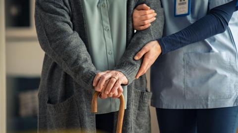 A care worker is holding a patients arm. The patient has both there hands on top off a wooden stick. The care worker is dressed in blue. The patient is wearing a green shirt with grey cardigan. 