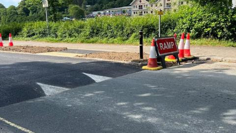 A table-top speed hump which forms part of a crossing installed on Malpas Road in Truro. Several traffic cones and a red warning sign saying ramp are on one side of the road. Buildings can be seen in the background past a hedgerow.