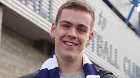 Will Kilgannon in a blue and white striped football scarf. He is standing in front of Millwall Football Club's home ground.