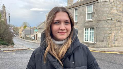 Anastasiia standing in the centre of the shot looking at the camera with the street and residential buildings on the background.