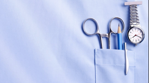 The picture shows the front of a light blue nurse uniform top. There is a pocket on the right-hand side of the image, and several items are tucked into it, a pair of metal medical scissors with rounded ends, a short pencil and a pen. Clipped to the top of the pocket there is a small fob watch with a silver strap and a white clock face.