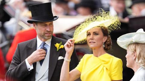 The Prince and Princess of Wales, William and Catherine, are pictured among crowds at Royal Ascot. Prince William, stood to the left, is wearing a three piece suit with a top hat. Princess Catherine is wearing a yellow dress and hat, the brim of which she is holding with her right hand. 