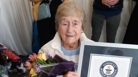 An elderly woman smiling at the camera. She is wearing a white fleece and has short blonde hair, while holding a silver photo frame with the Guinness World Records logo in it. The woman is also holding a bunch of flowers