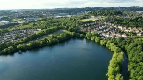 An aerial shot of a reservoir, it is surrounded by trees and a housing estate is nearby