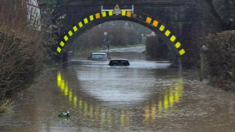 Severe flooding under Lockerley Rail bridge. Two cars are almost entirely submerged under the water. A green food waste caddy is seen floating by. 