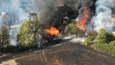 An aerial view of a fire engulfing gardens, with trees either side and house nearby. There is a scorched field in the foreground.