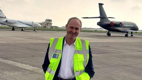 Steve Peters, smiling and wearing a high vis jacket, standing on the airport concourse with a small white plane behind him to the left and a grey one to the right. The airport buildings are in the distance