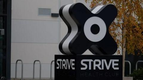 The outside of the Strive health and fitness centre in St Peter, Jersey. The large 'x' shaped branded Strive sign is visible in the foreground. A grey building is visible in the background. 