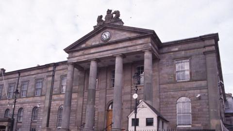 A close-up of Omagh courthouse on a cloudy day.  The large, grey stone building has arched Georgian windows and a Palladian portico with a large clock. 