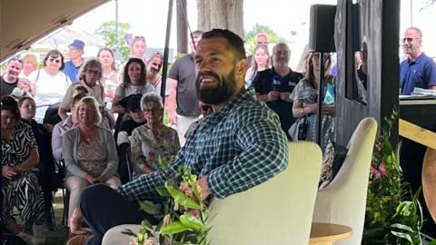 A man with short brown hair with shorter sides and a brown beard is smiling to the side. He is wearing a chequered blue shirt and sat on a beige seat on a stage. Behind him are several people sat in front of the stage on chairs and stood up at the back.