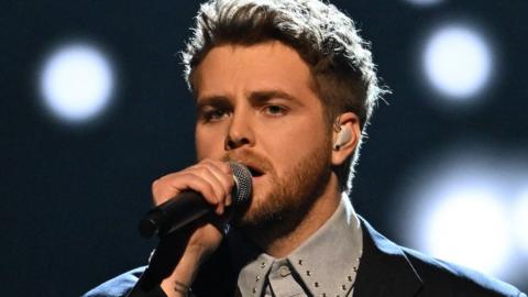 Singer-songwriter Alex Warren holds his microphone as he performs at the American Music Awards. Stage lights shine behind him.