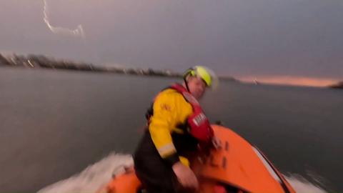 A man in an orange life boat is looking over his shoulder, towards the camera. He is wearing a yellow jacket and a red life jacket. The back ground is a mixture of water and cloudy skies with a faint tinge of orange as the sun sets.