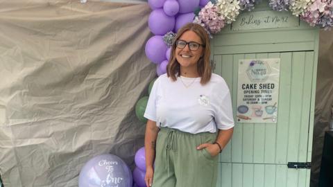A women with short brunette hair. She is wearing a white top and green trousers  whilst looking at the camera smiling. she is stood in front of a shed also green which is used to store her baked goods.