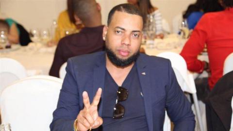 A man wearing a navy blazer with a navy T-shirt beneath it, which he has attached sunglasses to the neckline to, gives a "peace" sign with two fingers. He is sat at a table at what appears to be a wedding, with white cloth-covered seats and people surrounding a table behind him, drinking wine.