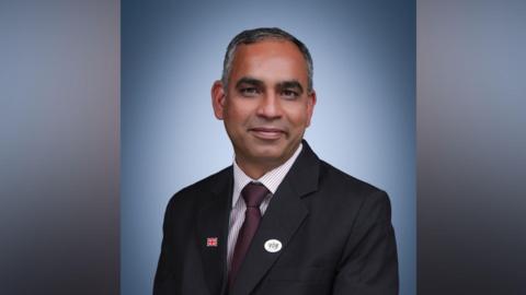 Adi Malviya wears a dark-coloured suit, striped shirt and purple tie. He faces the camera. He sits against a plain blue background.