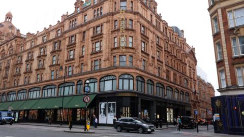 An exterior of Harrods department store. Traffic drives in front of the building.