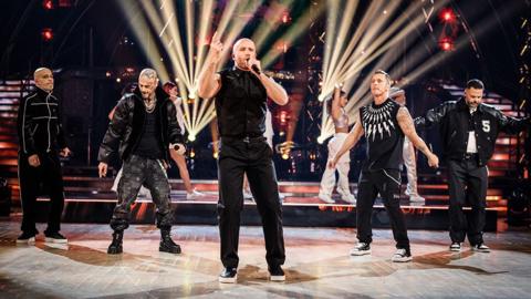 Five singing on the set of Strictly Come Dancing. They are all wearing black and dancing in front of white spotlights