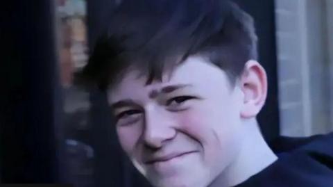 A boy with brow hair, wearing a navy hoodie, smiles at the camera, standing outside.