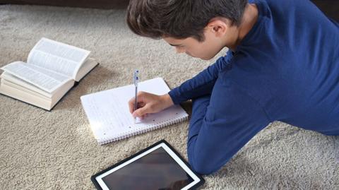 GCSE physics image: a teenager sits on the floor studying with a tablet, textbook and nodepad.