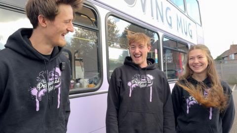 Josh Jones, Toby Jones and Maddy Harris wearing black hoodies that say "professional idiots" laugh in front of the lilac coloured bus they've converted into a coffee shop