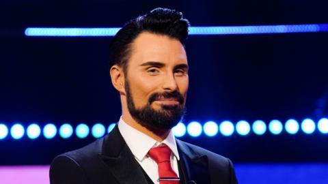 Rylan Clark, who is wearing a navy suit, red tie and smiling at the camera.