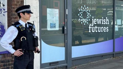 A policeman stands outside a building, with 'Jewish Futures' written on the glass panel