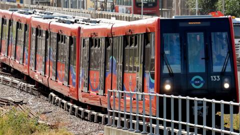Red and blue Docklands Light Railway train on tracks behind a metal barrier. The front of the carriage is black with a DLR roundel