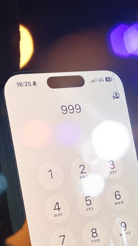 A mobile phone with 999 typed in with flashing lights in the background.