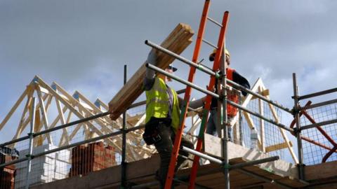 A pair of roofers work on a housing construction site. The timber structure of a roof can be seen and one man is holding planks on his shoulder and talking to the other man. Scaffolding, ladders and railings are being used.
