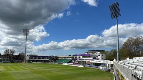 The Essex County Cricket Ground on a sunny day. The lawn and stands are visible, with a blue sky and cloud above.
