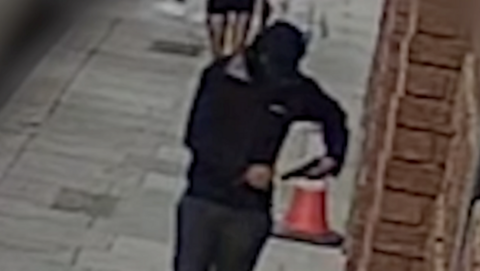 A hooded man dressed in black carries a gun in the street