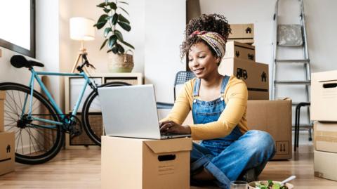 A young woman sitting on the floor in dungarees, smiling and wearing a head wrap to hold up her hair, she is working on a laptop which is propped up on a cardboard box - behind her there are a number of boxes and a ladder is propped against a wall, showing someone who has just moved 