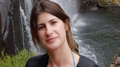 Woman with brown hair tied back looking at the camera with her head to one side - and a waterfall seen behind.