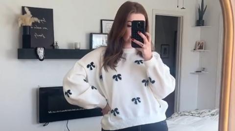 A woman in a white jumper with bows on it looks in the mirror holding her phone