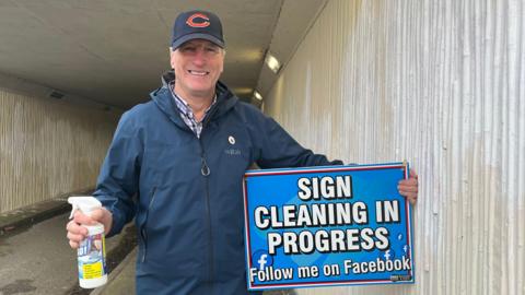 Photograph of John, who is looking into the camera and smiling. He is wearing a dark blue raincoat, with a purple checked shirt underneath. He is also wearing a dark blue cap with an orange letter C on it. He is holding a blue sign, that reads in white letters 'Sign Cleaning in Progress, follow me on Facebook', and in the other hand has a white spray bottle. He is standing in the underpass, which has cream coloured, corrugated walls which have large patches of white paint where he has begun to paint over the graffiti. 