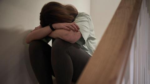 A woman sits on the stairs of her home with her head in her arms, covering her face