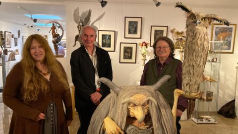 Wendy Froud, Alan Lee, and Brain Froud stand amongst their art in the Green Hill Arts gallery. They are gathered around a large model of the Troll Witch, one of Wendy's creations. 