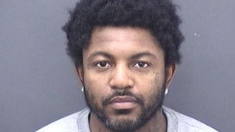 A police custody photo against a grey background shows Andre Willis with an Afro hairstyle and a short beard. He wears earrings and a light, collarless top.