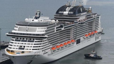 A white cruise ship with multiple decks and flanked by tug boats on calm water.