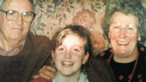 Katie pictured as a child, with blond hair and red sweatshirt. She's sitting between her grandfather and grandmother on the sofa
