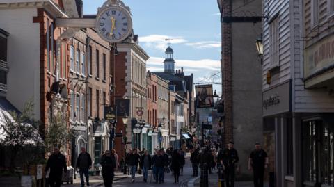 Rochester High Street on a sunny day, with many people walking along streets lined with historic brown and red brick buildings.